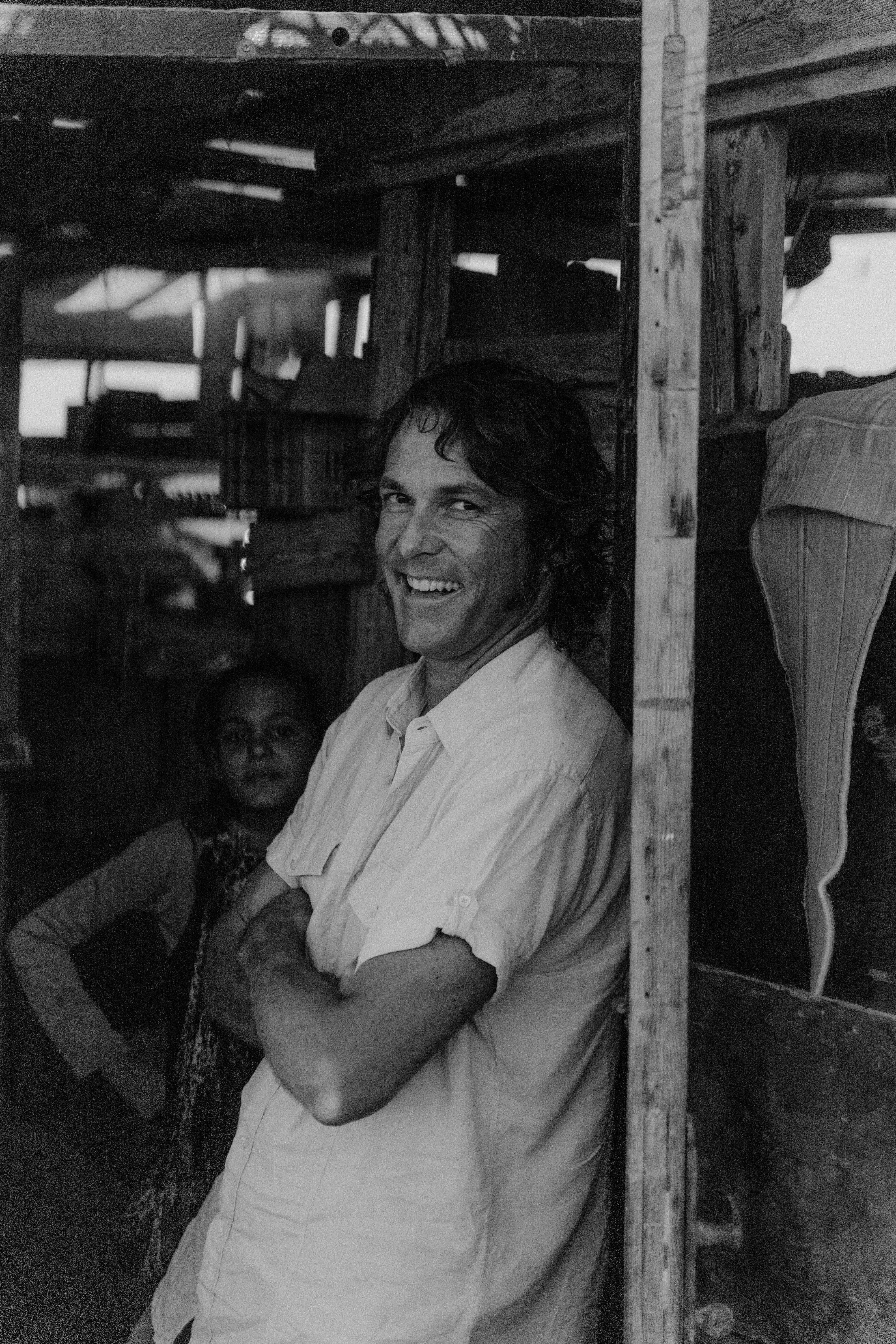 grayscale photo of a man in button up shirt leaning on a wooden post