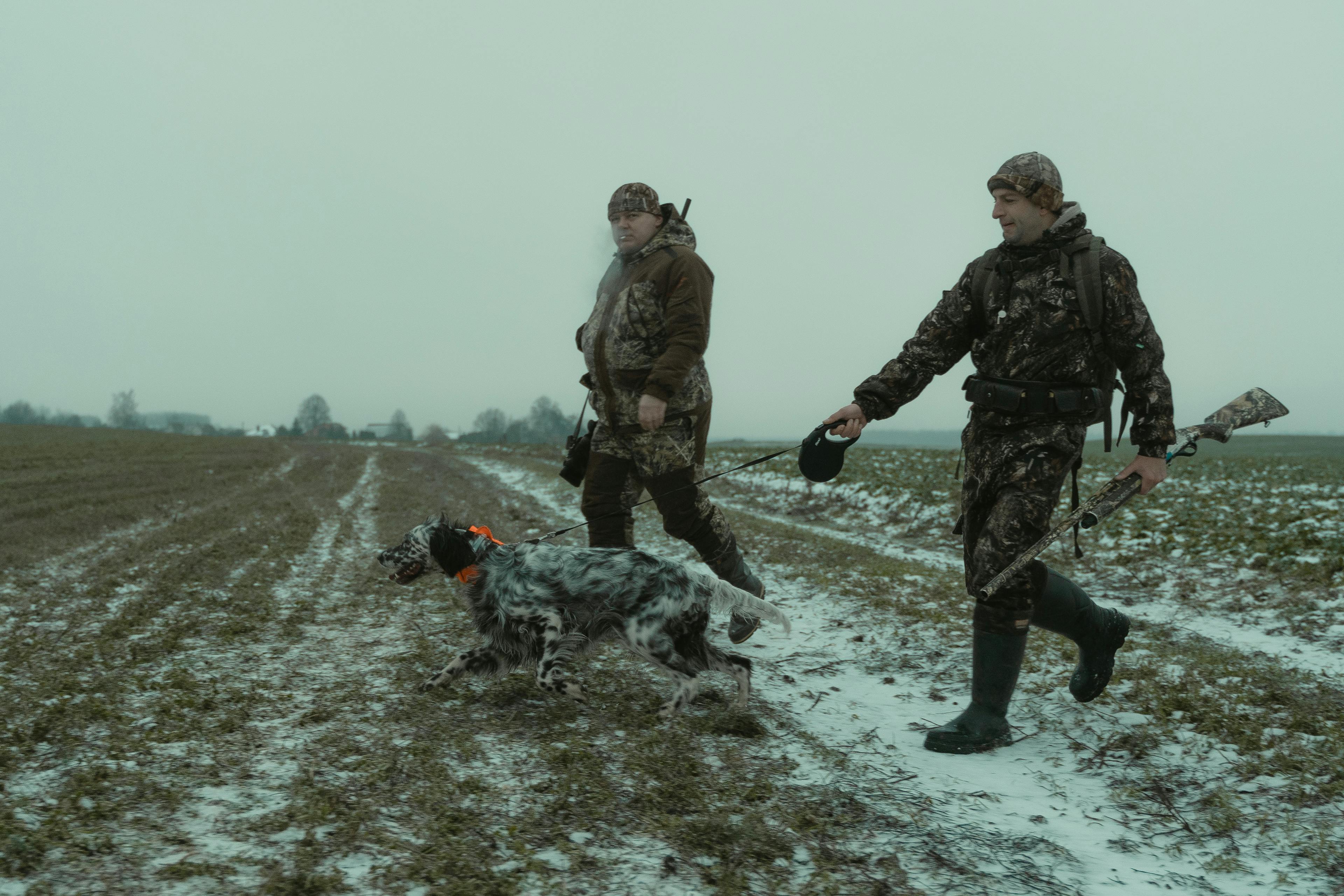 two men in camouflage walking in the field with a dog